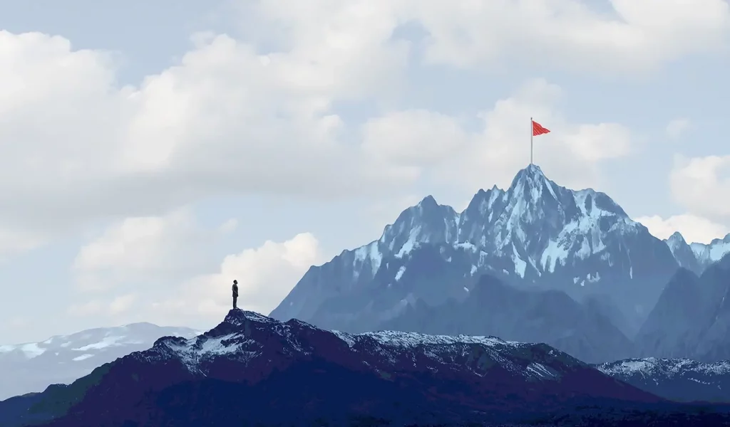 A man standing far from a mountain and the mountain has a red flag on it in the distance